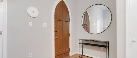 A hallway with a wooden door, a round mirror, a black console table, and a clock on the wall.