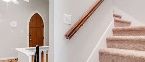 Un escalier avec des marches recouvertes de moquette et une rampe en bois, un mur blanc orné d’une horloge, et une porte en bois surmontée d’un arc.