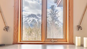 Quarto com uma grande janela com vista para uma paisagem montanhosa coberta de neve. Há um banco de madeira e uma pequena mesa com velas.
