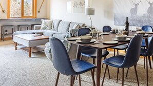 Uma área de jantar com uma mesa de madeira posta para seis pessoas, rodeada por cadeiras azuis. Um sofá cinza e uma pequena mesa de centro estão ao fundo.