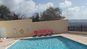 A rectangular swimming pool with clear blue water, surrounded by a beige tiled deck. There are four red lounge chairs aligned along the poolside. A lifebuoy is attached to the wall, and a sign indicates the pool is private. - 22C Christina Hilltop Apartment panoramic views. (Peyia)