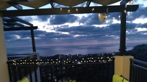 An outdoor patio with a table and chairs, string lights, and a view of the city at dusk. - 22C Christina Hilltop Apartment panoramic views. (Peyia)
