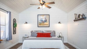 A bedroom with a sloped ceiling, a bed with a white comforter and red throw pillows, a framed rooster picture above the bed, and a small table with a lamp beside the bed.
