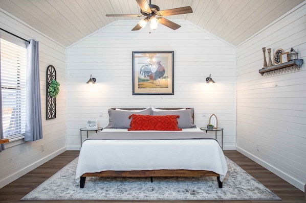 Un dormitorio con techo inclinado, una cama con cubrecama blanco y almohadones rojos, un cuadro de gallo enmarcado sobre la cama, y una mesita de noche con una lámpara a un lado.