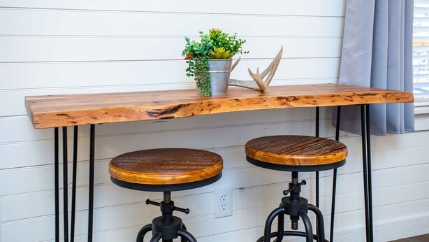 A wooden bar stool with a round seat and a metal base, positioned under a wooden table with a metal top.