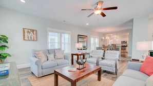A spacious living room with a light blue sofa, a wooden coffee table, and patterned armchairs. A ceiling fan and a kitchen are visible in the background. - Warm Waters Oasis at Devonshire Place (Kill Devil Hills)
