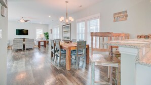 A spacious dining area with a wooden table and chairs, a chandelier, and a television mounted on the wall. - Warm Waters Oasis at Devonshire Place (Kill Devil Hills)