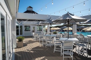 An outdoor patio with white tables and chairs, umbrellas, and a pool in the background. - Warm Waters Oasis at Devonshire Place (Kill Devil Hills)