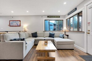 Una sala de estar moderna con un sofá modular, una mesa de centro de madera y un ventanal con vistas a una casa.