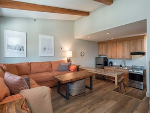 A cozy living room with a sofa, a wooden coffee table, and a kitchen area with wooden cabinets and a stove.