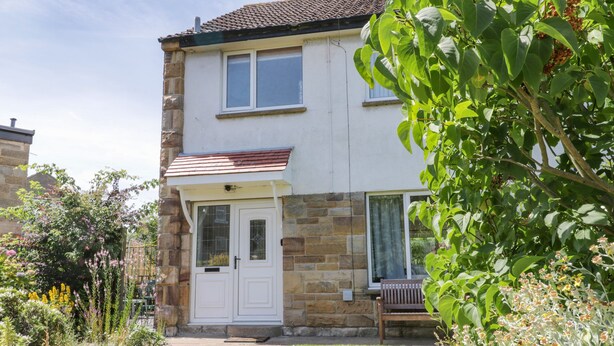 A two-story house with a stone facade, a white door, and a small porch with a bench. - Sea Change (Whitby)