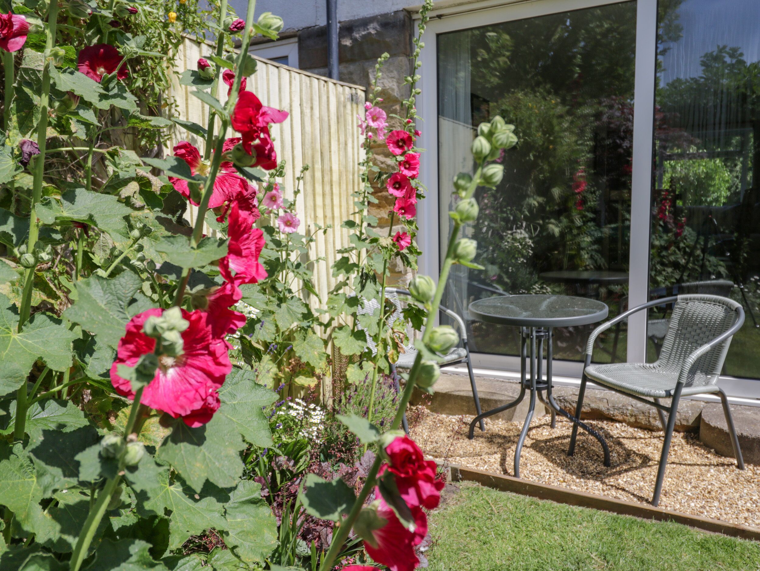 Un jardín con flores rojas, una puerta de cristal y una silla de metal.