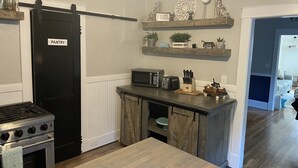 A kitchen with a stove, microwave, toaster, and wooden cabinets. There is a 'PANTRY' sign on the door, and shelves with decorative items above the counter.