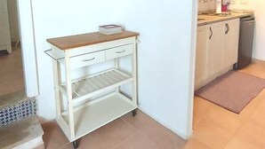 A kitchen with white cabinets, a wooden countertop, and a small white shelf unit. There is a white air conditioning unit on the wall, and two framed pictures above the shelf.