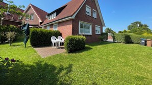 A red brick house with a sloped roof, a paved driveway, and a small garden area with a white chair and table. - Strandläufer (Wangerland)