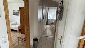 A hallway with a bed, a small table, a trash bin, and a door leading to another room. - Strandläufer (Wangerland)