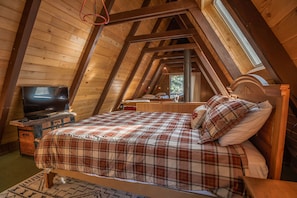 A cozy attic bedroom with a wooden ceiling, a bed with plaid bedding, a television on a wooden dresser, and a window providing natural light.