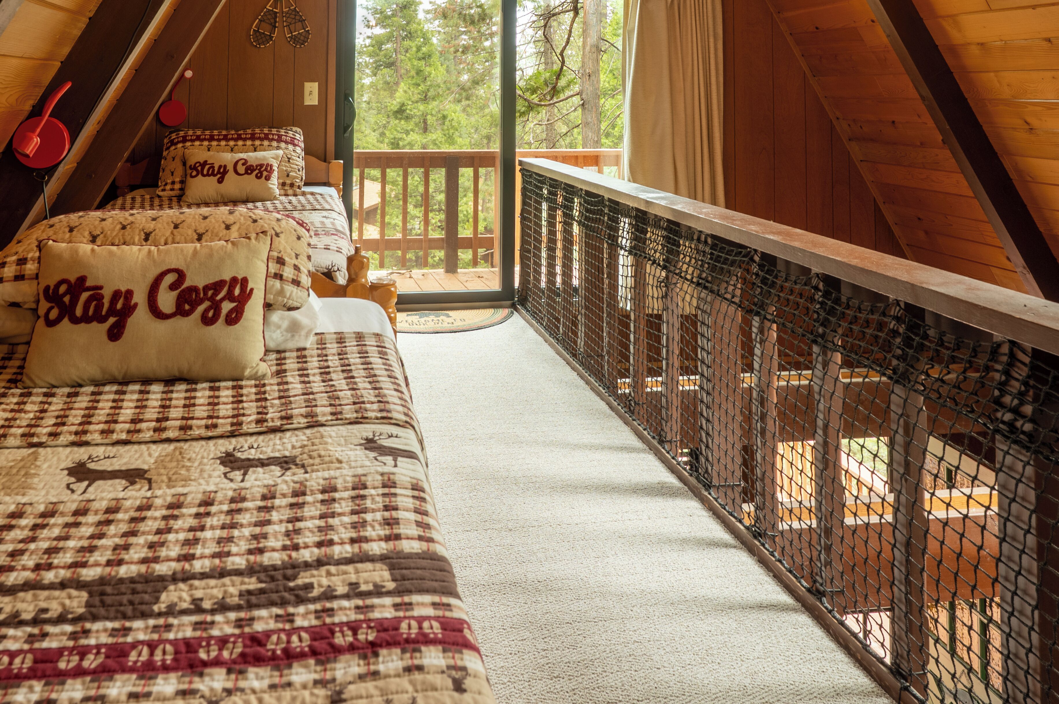 Una habitación acogedora con una colcha a cuadros, dos almohadas con el texto 'Stay Cozy', vista al exterior a través de un ventanal y barandilla de balcón de madera.