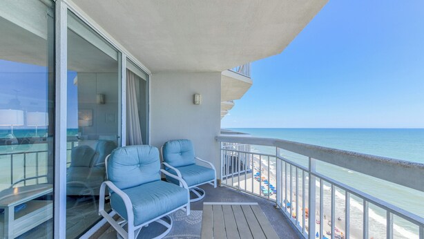 A balcony with two blue cushioned chairs, a glass door leading to a room, and a view of the beach with umbrellas.