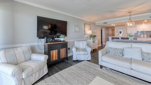 A living room with a sofa, armchairs, a TV, and a fireplace. There is a kitchen in the background with a dining area.