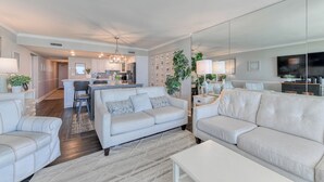 A spacious living room with a sofa, a coffee table, and a TV. There is a kitchen area in the background with a dining table and chairs.