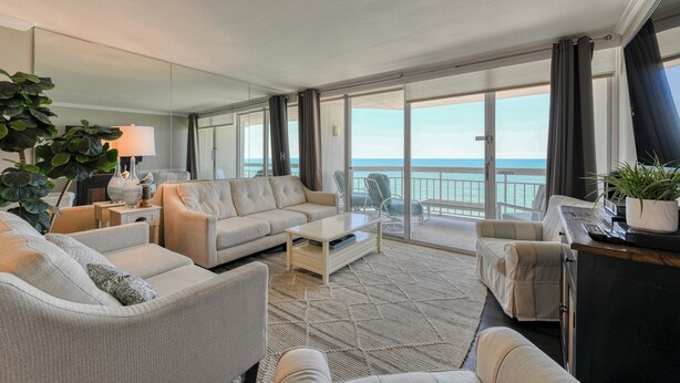 A spacious living room with a large sofa, a coffee table, and a view of the ocean through large sliding glass doors.