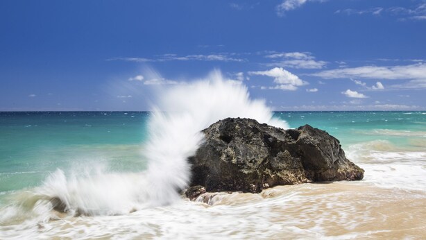 Ein felsiger Felsen, an dem Wellen brechen, ein klarer blauer Himmel und ruhiges Meer.