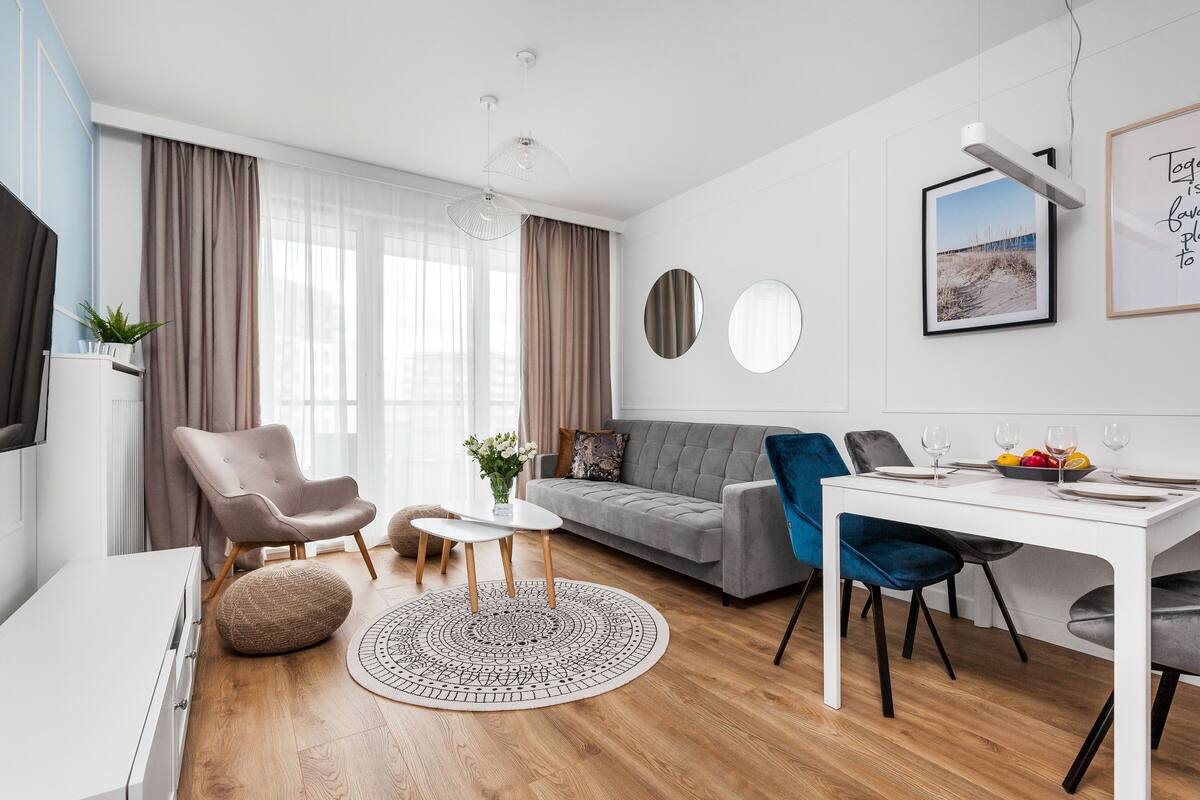 Una sala de estar moderna con un sofá gris, una mesa de comedor blanca y una silla azul. Hay un televisor a la izquierda y una ventana con cortinas.