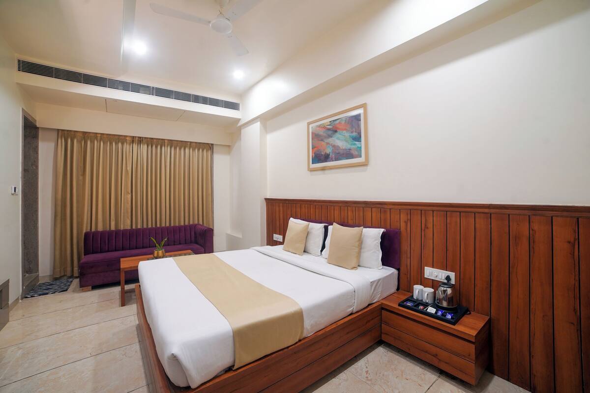 Una camera d'albergo con un letto grande, una testiera in legno, un tavolino con un vassoio di oggetti, un divano viola e un quadro appeso al muro.