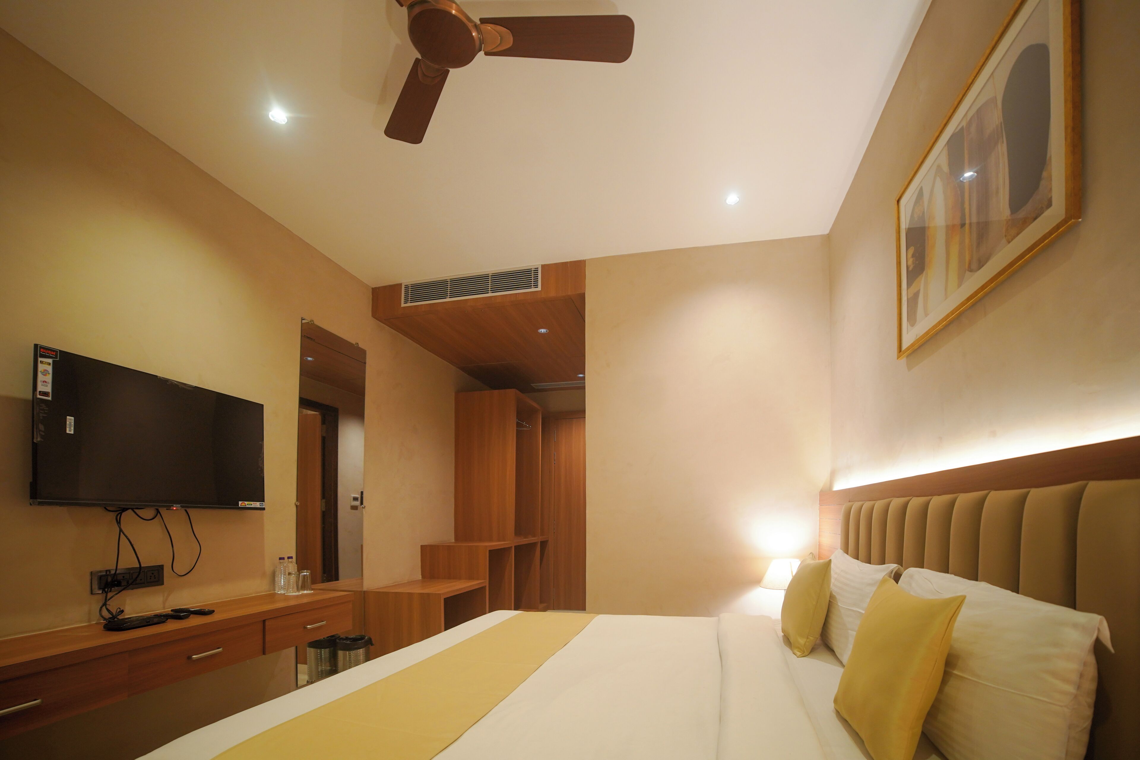 A hotel room with a bed, a television, a ceiling fan, a wardrobe, and a painting on the wall.