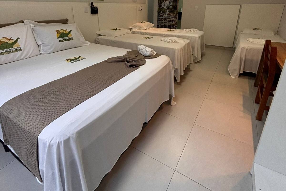 Habitación de hotel con dos camas individuales, una cama grande y una mesita. Hay un cuadro en la pared y una ventana con vistas al exterior.
