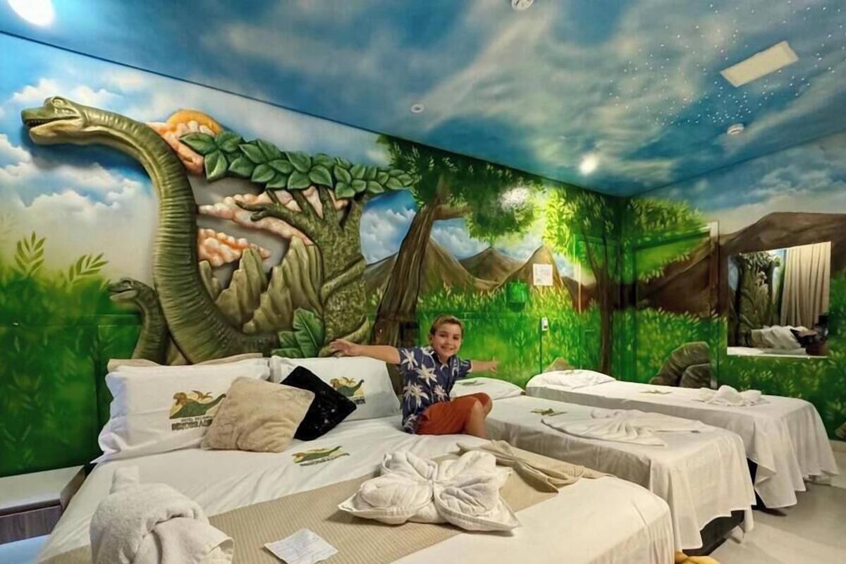 Habitación de hotel con un mural con temática de dinosaurios, varias camas y un niño sentado en una de ellas.