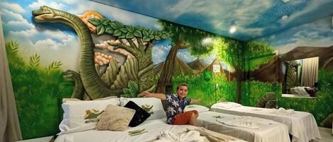 Habitación de hotel con un mural con temática de dinosaurios, varias camas y un niño sentado en una de ellas.