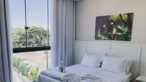 Habitación de hotel con un ventanal grande, una cama con ropa de cama blanca, un cuadro de dinosaurio y una mesita de noche pequeña.