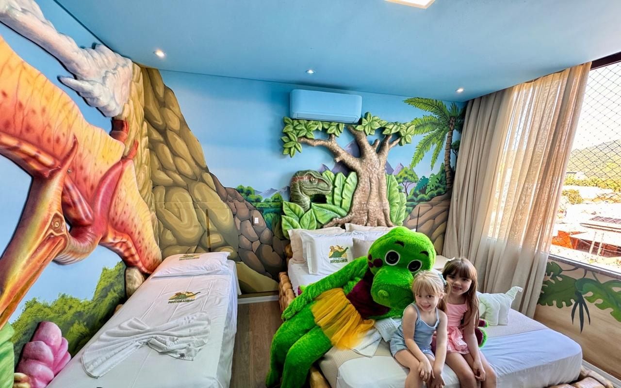 Een kamer met een muurschildering over dinosaurussen, een bed en een groot pluche dinosaurus speelgoed.