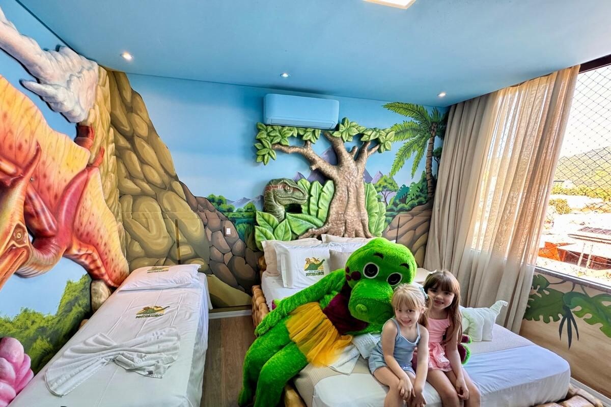 Una habitación con un mural con temática de dinosaurios, una cama y un gran peluche de dinosaurio.