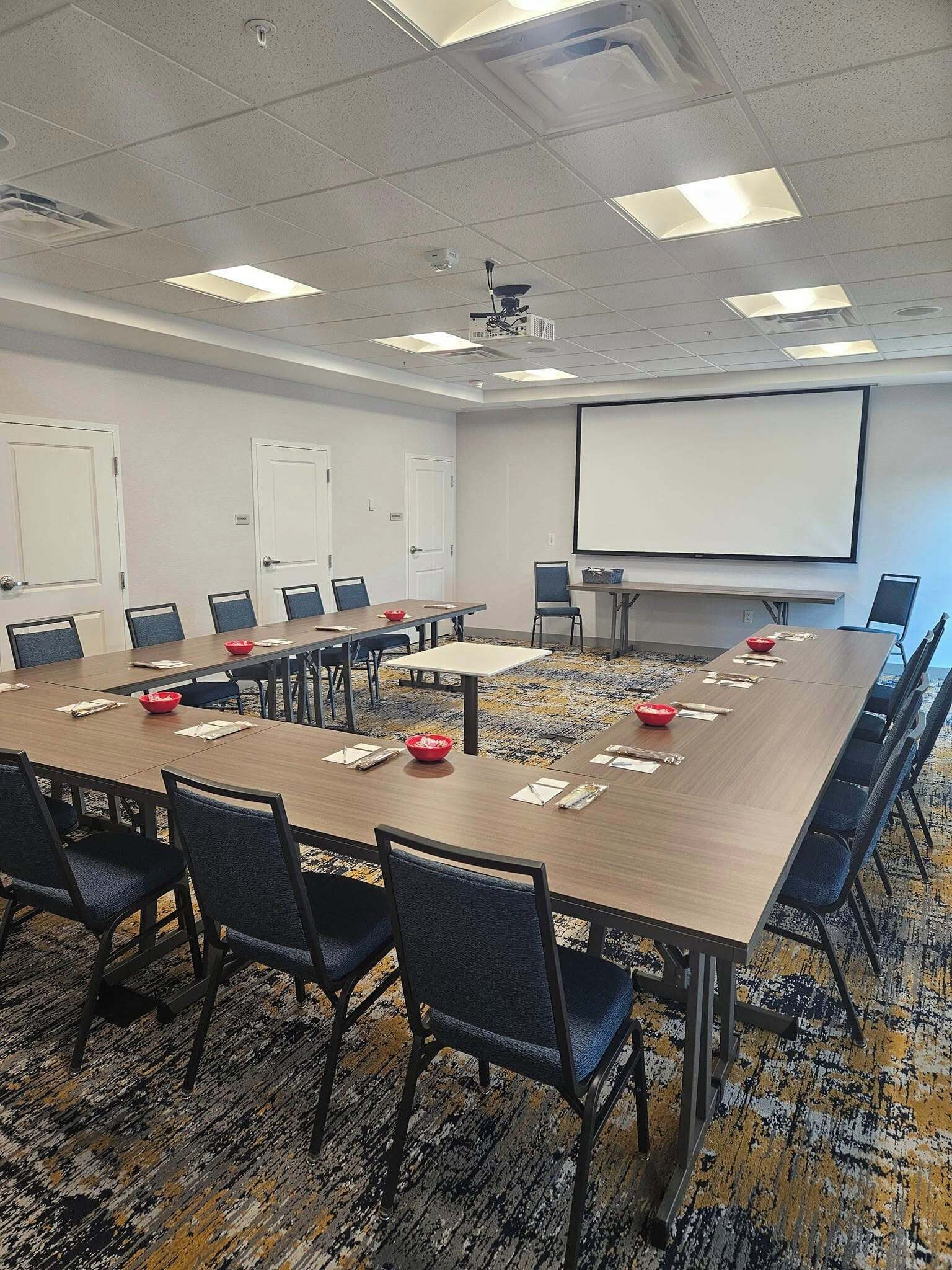 A conference room with a long table, chairs, a projector screen, and a carpeted floor.