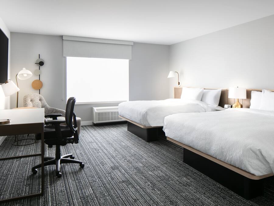 A hotel room with two beds, a desk, a chair, a TV, and a large window.