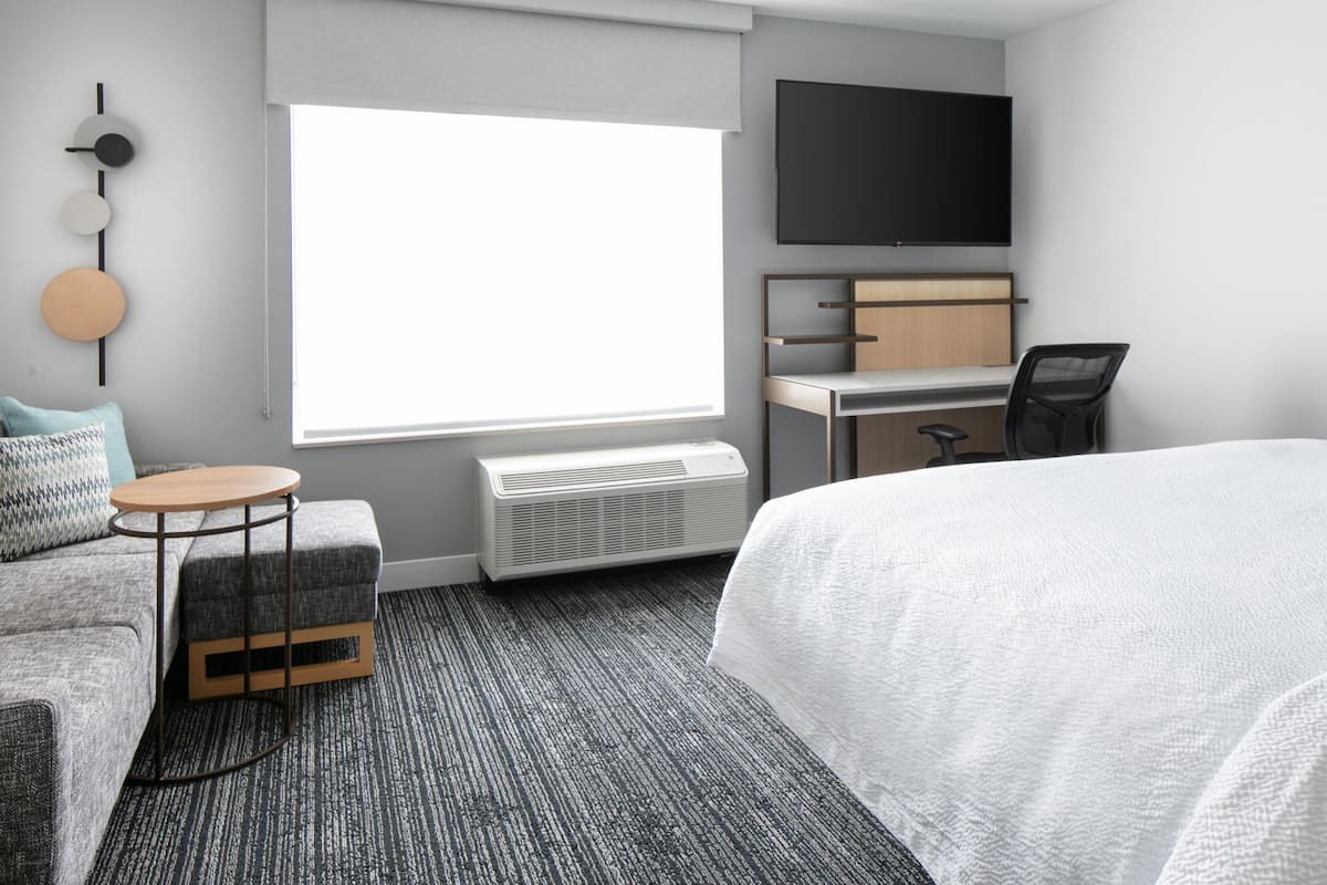 Een moderne hotelkamer met een groot bed, een bureau met stoel, een ronde tafel, een tv aan de muur en een groot raam met jaloezieën.
