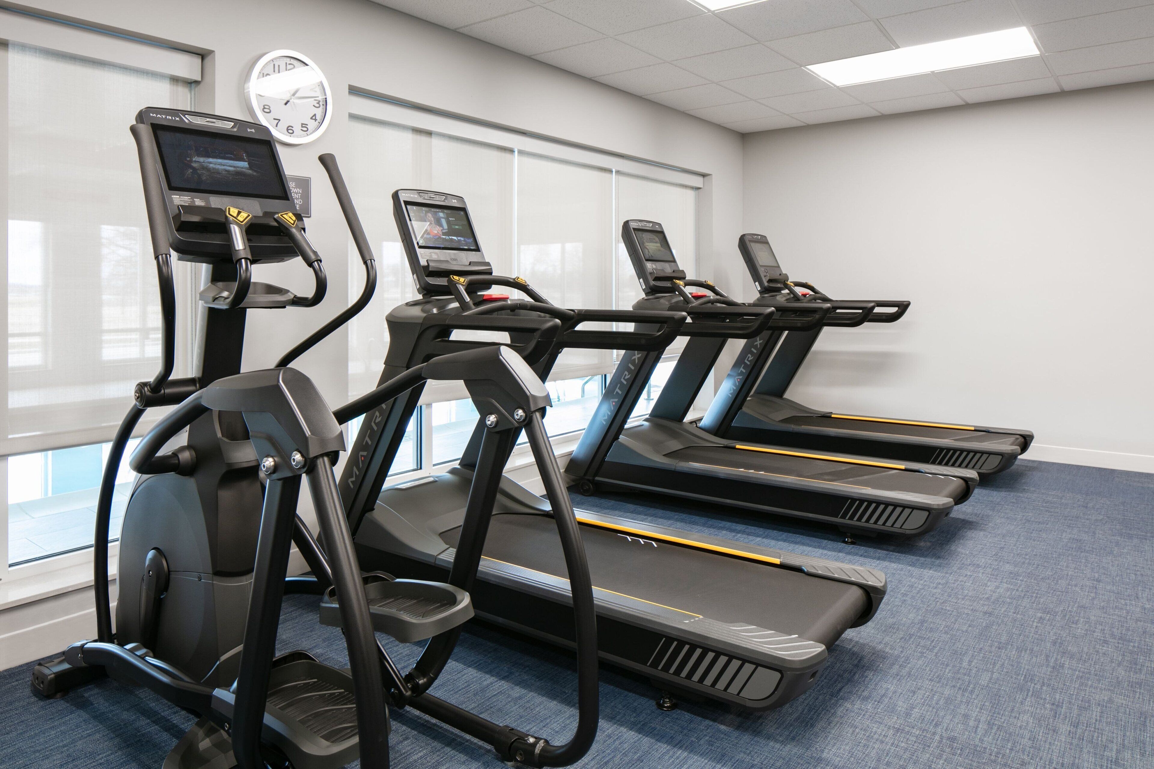 A fitness room with treadmills and a stationary bike, large windows, and a clock on the wall.