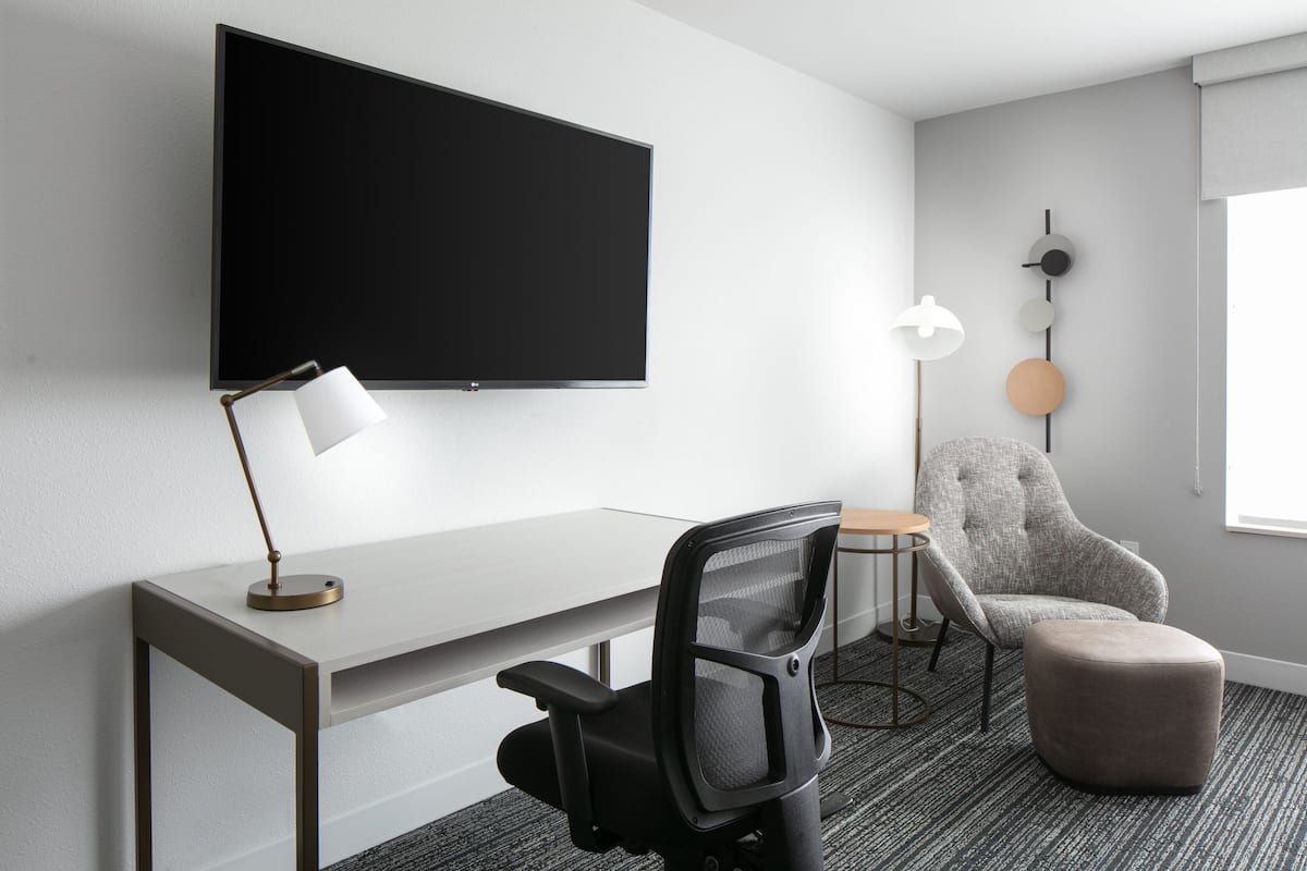 Een moderne hotelkamer met een flatscreen televisie aan de muur, een bureau met een lamp, een fauteuil en een rond hocker.