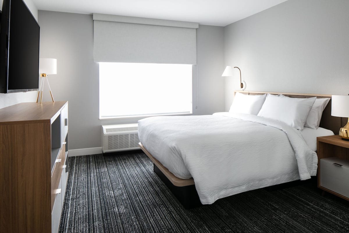 Een hotelkamer met een bed, nachtkastje, lamp en een aan de muur bevestigde televisie.