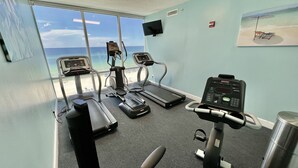 A fitness room with treadmills, a stationary bike, and a rowing machine, overlooking a beach. - Beach-Front Condo, 2 Reserved Parking Spaces!, Large Balcony, Fabulous Views! (Panama City Beach)