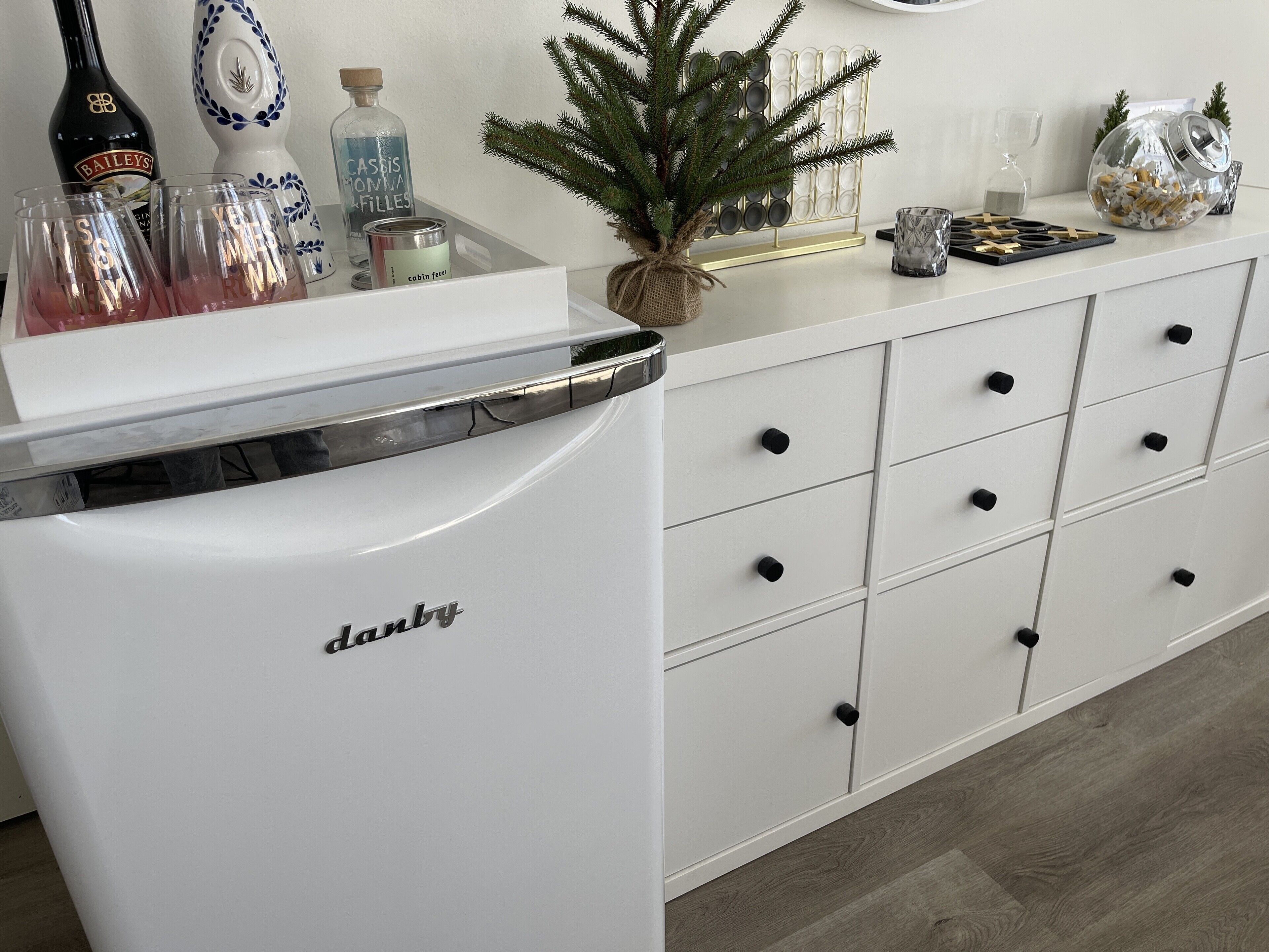 Un refrigerador blanco con el nombre de marca 'danny', un mueble blanco con varios cajones y una pequeña planta en maceta encima.