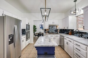 A modern kitchen with white cabinets, a stainless steel refrigerator, a marble island, and a pendant light.