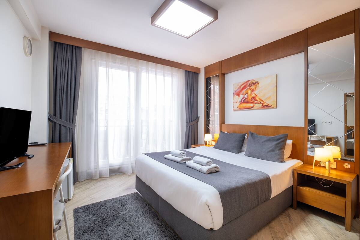A modern hotel room with a large bed, a desk with a computer, a TV, and a large window with curtains.