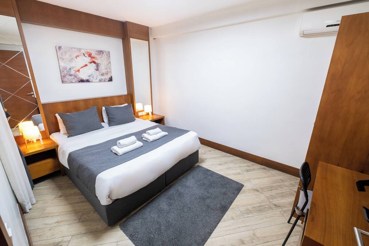 A modern hotel room with a large bed, a nightstand with a lamp, a painting on the wall, and a wooden wardrobe.