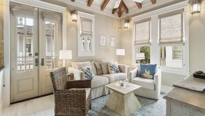 Ein geräumiges Wohnzimmer mit hoher Decke, sichtbaren Holzbalken und großen Fenstern mit Jalousien. Es gibt zwei Sofas, einen Korbstuhl und einen Couchtisch.