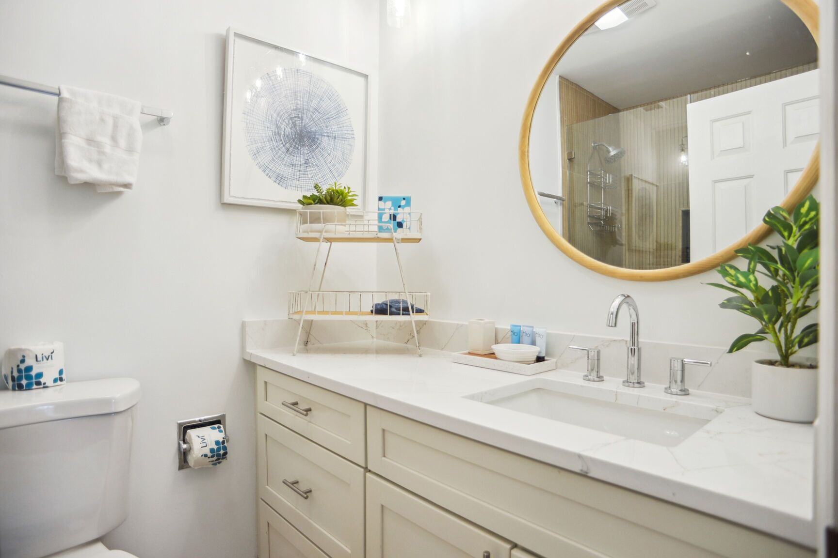 A modern bathroom with a white sink, vanity, and toilet. There is a framed artwork above the sink, a small shelf with plants, and a round mirror with a gold frame.