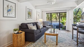 A modern living room with a dark grey sofa, a glass coffee table, and a patterned rug. There are framed artworks on the walls and a view of a lush garden through large sliding doors.